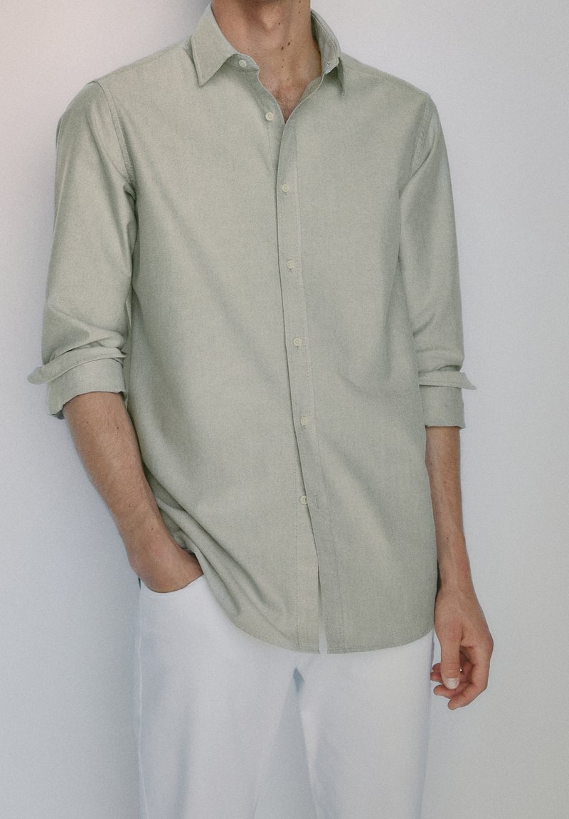 Homme portant une chemise boutonnée vert clair avec manches retroussées et un pantalon blanc, debout avec une main dans la poche devant un mur uni.