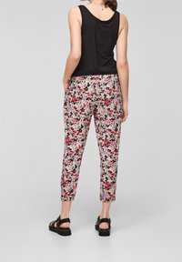 Femme portant un haut noir sans manches et un pantalon court à motif floral avec des sandales noires, debout devant un fond gris uni.