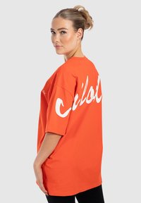 Oranžové oversized tričko s krátkými rukávy, s velkým bílým textem na zádech. Vyrobeno z jemného bavlněného materiálu, s volným střihem.