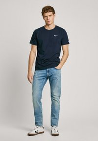 Navy blue cotton t-shirt with short sleeves, round neckline, and small logo on the chest, paired with light blue jeans and white sneakers.