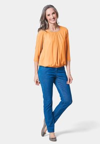 Oranges, plissiertes Oberteil mit Dreiviertelärmeln, kombiniert mit blauen, schmal geschnittenen Hosen. Graue Ballerinas mit Schleifenakzent. Modell posiert vor einem weißen Hintergrund.