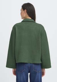 Grøn, tekstureret, oversized jakke med krave og brede ærmer, parret med blå denimjeans. Synlige sømme og et cropped design.