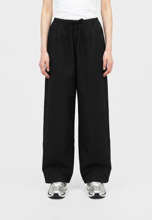 Personne portant un pantalon noir ample avec cordon de serrage et des chaussures de sport argentées, debout devant un fond clair uni.