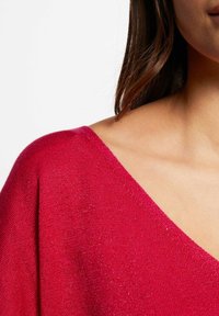 Rode gebreide trui met een V-hals ontwerp, met een zachte textuur en subtiele metallic draadaccenten. Close-up van de schouder en de halslijn.