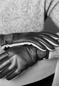 Guanti in pelle nera con accenti bianchi. Presentano una toppa rotonda con logo. Design elegante e taglio aderente. Sfondo in morbida lana.