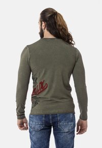 Olivgrünes Langarm-T-Shirt mit grafischem Druck in Rot auf der Rückseite. Weicher Stoff, taillierter Schnitt und Rundhalsausschnitt. Sichtbare Jeans aus Denim.