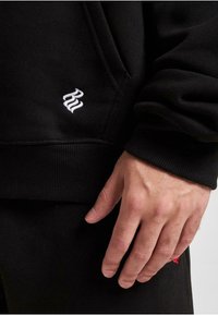 Zwarte hoodie gemaakt van een zachte katoenmix, met een witte geborduurde logo op de zak en ribbelmanchetten en -zoom. Hand weergegeven voor schaling.