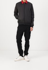 Černá bomber bunda s červeným límcem, zapínáním na zip a žebrovanými manžetami, v kombinaci s černými cargo kalhotami a bílými teniskami. Minimalistický design.