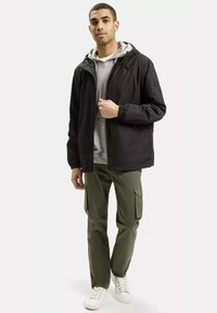 Czarna lekka kurtka z kapturem, z elastycznymi mankietami, noszona na szarym hoodie; połączona z oliwkowymi spodniami cargo i białymi trampkami.