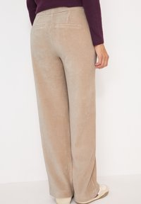 Persoon draagt beige corduroy wijde broek en crèmekleurige sneakers, met een zichtbaar paarse mouw van een trui en hand aan de zijkant.