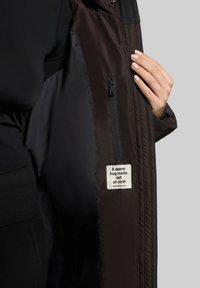 Veste noire matelassée avec un tissu extérieur lisse, poche zippée sur la doublure intérieure et une étiquette indiquant "Un câlin chaud en tissu."