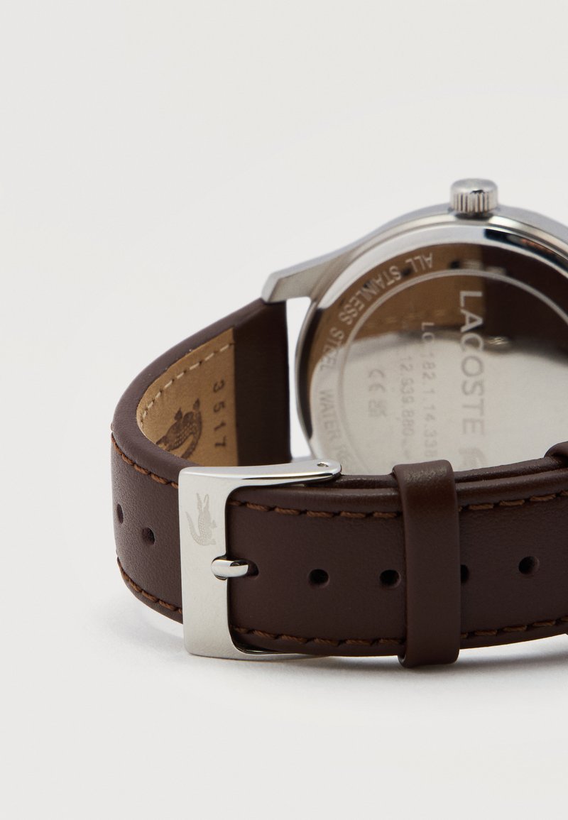 Braunes Lederarmband mit silberner Schnalle und geprägtigem Krokodillogo, befestigt an einem silbernen Gehäuseboden einer Lacoste-Uhr.