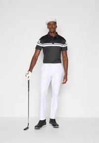 Μαύρο πόλο μπλουζάκι με λευκές ρίγες, λευκά παντελόνια, μαύρα γκολφ παπούτσια και λευκό καπέλο. Κράτα μια γκόλφ κλάμπ στο χέρι, σε στάση, με φόντο άσπρο.