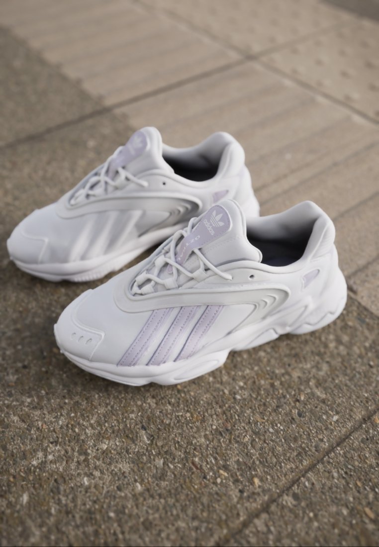 Scarpe sportive bianche con accenti viola. Realizzate in materiale sintetico liscio, presentano lacci, etichetta con logo e un design di suola sagomata.