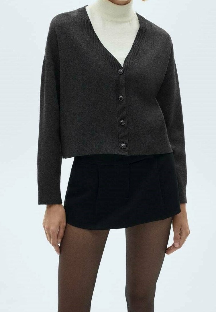 Femme portant un cardigan noir court boutonné sur un col roulé blanc, une mini-jupe noire et des collants noirs transparents, debout devant un fond uni.