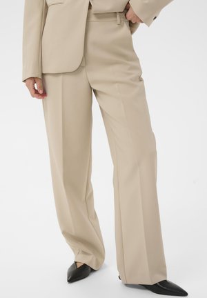 Beige maßgeschneiderte weit geschnittene Hosen, kombiniert mit einem passenden Blazer und schwarzen spitzen Schuhen vor einem weißen Hintergrund.