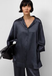 Camicia oversize di un blu scuro navy con un tessuto texturizzato e lucido, chiusura a bottoni e colletto ampio. Abbinata a una borsa nera.