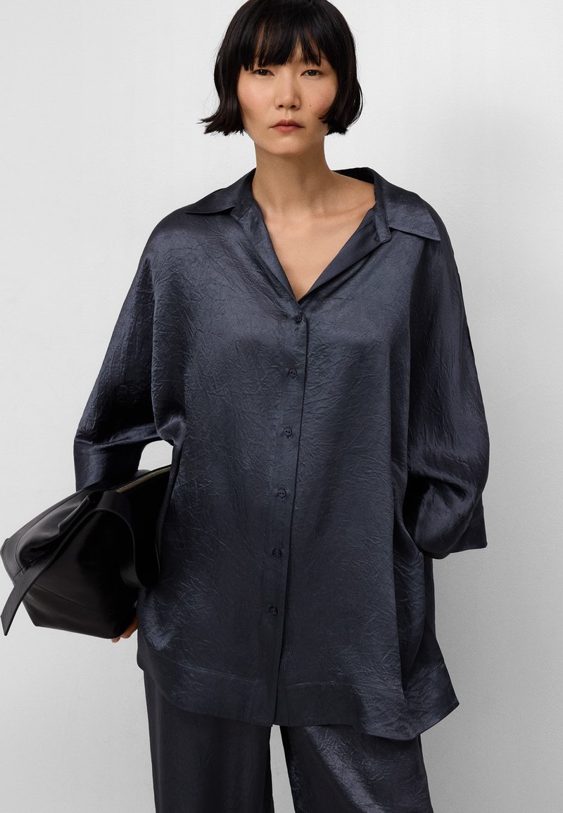 Camicia oversize di un blu scuro navy con un tessuto texturizzato e lucido, chiusura a bottoni e colletto ampio. Abbinata a una borsa nera.