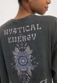 Žena nosi tamnosivu majicu s natpisom „Mistična energija“ i plavim cvjetnim i listnim dizajnom na gornjem dijelu leđa.