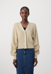 Beige strikket cardigan med v-hals, fem knapper og ribbestrikket tekstur. Har puffermer og en cropped design, sammen med et denimskjørt.
