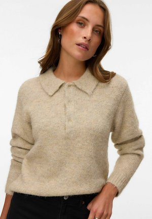 Jeune femme aux cheveux châtain clair portant un pull en tricot beige avec col et boutons et un pantalon noir, posant avec une main dans la poche.
