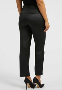Pantalon noir sur mesure avec un motif croco texturé, présentant une coupe slim, une ceinture et des poches arrière, associé à des stilettos noirs étincelants.