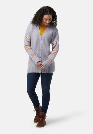 Lichtpaarse cardigan van textiel, met v-hals en knopen aan de voorkant. Accentuated met gebogen mouwen in oranje en geel. Gecombineerd met blauwe jeans en bruine laarzen.