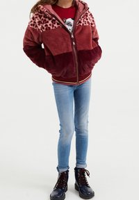 Faux fur-jacket i röda och rosa nyanser, med leopardmönstrad ovan, dragkedjestängning, ribbade ärmslut och nederkant. Kombinerad med blåjeans och svarta stövlar.