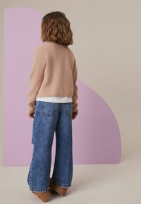 Bež pleteno pulover, pod njim bela srajca, v kombinaciji s širokimi modrimi džinsami. Ozadje s purpurnim abstraktnim oblikom.