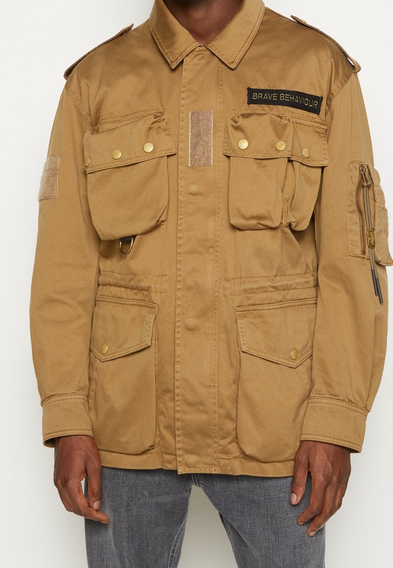 Beżowa kurtka typu utility z wieloma przednimi kieszeniami, złotymi zatrzaskami, naszywką na piersi oraz zamykaną na zamek kieszenią na rękawie. Tkanina o fakturze.
