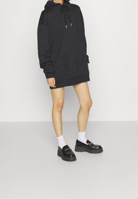 Vestido sudadera negro de gran tamaño con capucha ajustable y bolsillo frontal, combinado con calcetines blancos y mocasines negros de plataforma. Textura de tela suave.