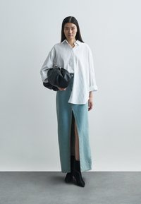 Biała koszula w cienkie prążki w stylu oversize, jasnoniebieska długa spódnica jeansowa z rozcięciem z przodu, czarna torebka oraz czarne botki na obcasie.