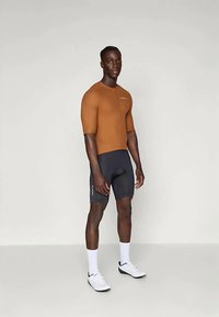 Syklistdrakt med kort erme i brun og svart med glidelås foran, med en smal passform og glatt tekstur, båret med hvite sko og sokker.