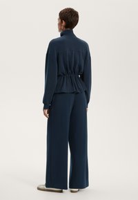 Mornarško modra, sproščena pulover z visokim ovratnikom in gubanjem v pasu, v kombinaciji s širokimi hlačami. Gladka tkanina, minimalističen dizajn.