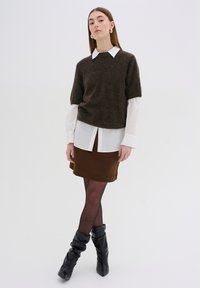 Pull en maille marron à manches courtes sur une chemise blanche à col, associé à une jupe en satin marron et des bottes noires plissées jusqu'aux genoux.