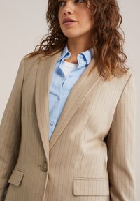 Smėlio spalvos dryžuotas švarkas iš tekstūruotos medžiagos, su viena sagos užsegimu ir išpjautomis atlapomis, derinamas su šviesiai mėlyna marškinėliais su sagomis.