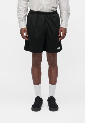 Mand iført sorte mesh-shorts med hvidt logo, hvide knælange sokker, sorte sneakers og en langærmet skjorte med striber, stående på hvid baggrund.