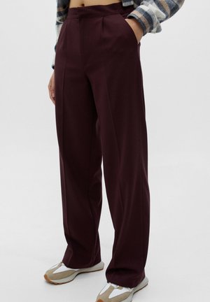 Personne portant un pantalon taille haute, jambes larges, de couleur bordeaux foncé, avec une main dans la poche, associé à des baskets décontractées beige et blanches.