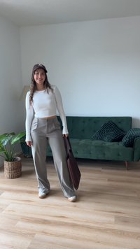 Brown cap, white long-sleeve top, gray high-waisted pants, and brown bag. Green couch with textured pillows and hardwood floor in background.