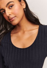 Ciemnogranatowy prążkowany top z dekoltem w kształcie litery U, z długimi rękawami i fakturowaną tkaniną. Minimalistyczny dodatek w postaci małego kolczyka w kształcie serca.