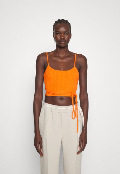 Πορτοκαλί κοντό τοπ με υφή, λεπτές τιράντες και δέσιμο στη μέση. Συνδυάζεται με ανοιχτό γκρι, κάπως χαλαρές παντελόνες.