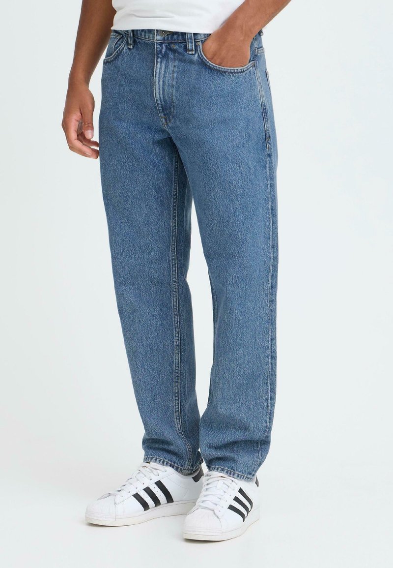 Mann iført blå rette jeans og hvite sneakers med svarte striper, en hånd i jeanslommen mot en ensfarget bakgrunn.