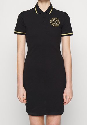 Kobieta nosi dopasowaną czarną sukienkę polo z złotym obszyciem na kołnierzu i rękawach oraz okrągłe logo Versace Jeans Couture na piersi.