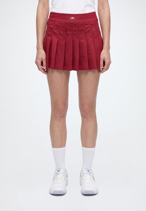 Un mannequin porte une jupe de tennis rouge plissée, un haut blanc, des chaussettes blanches à tige mi-mollet et des chaussures de tennis blanches.