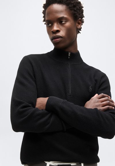 Νέος άνδρας με κοντά κυματιστά μαλλιά, φοράει μαύρο υφασμάτινο πουλόβερ με φερμουάρ στο μισό μήκος, έχει σταυρωμένα τα χέρια και κοιτάζει προς τα πλάγια με φόντο έναν απλό τοίχο.