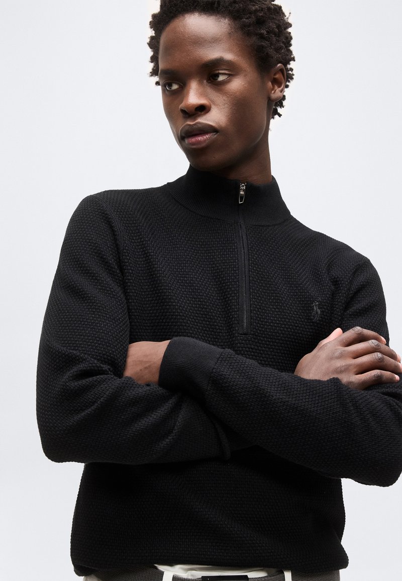 Jeune homme aux cheveux courts et bouclés portant un pull noir texturé à demi-fermeture éclair, bras croisés, regardant sur le côté devant un fond uni.