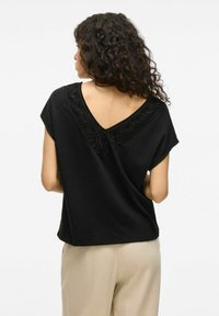 Top corto nero a maniche corte con scollatura a V e motivi circolari decorativi lungo il colletto. Il tessuto sembra leggero con una vestibilità rilassata.