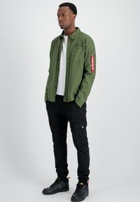 Camisa verde de botones con dos bolsillos en el pecho y un bolsillo con cremallera. Combinada con pantalones cargo negros que tienen bolsillos laterales. Zapatos negros.
