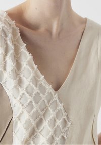 Vestido beige claro con escote en V, adornado con un panel texturizado en blanco roto decorado con un patrón de diamantes en un hombro.