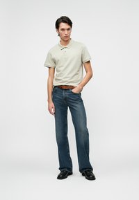 Světle béžová polokošile s krátkými rukávy, kombinovaná s modrými zvonovými džíny a hnědým koženým pásem. Černé boty, neutrální pozadí.